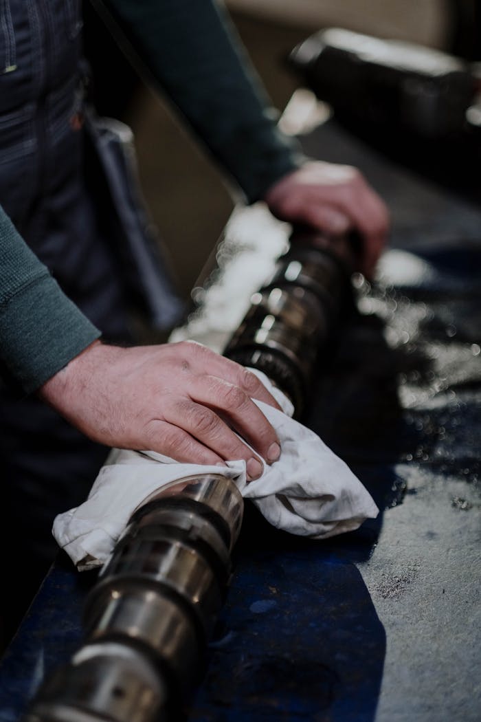 Hands cleaning a greasy camshaft with a cloth in a mechanical workshop.